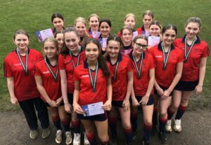 A group of girls are stood together holding certificates and wearing their medals.