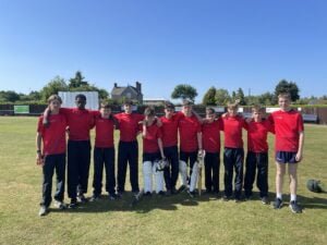 11 boys are stood together in a group outside on the cricket field.